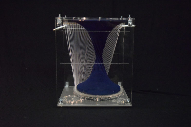 acrylic box holding navy textile form and band of strings inside