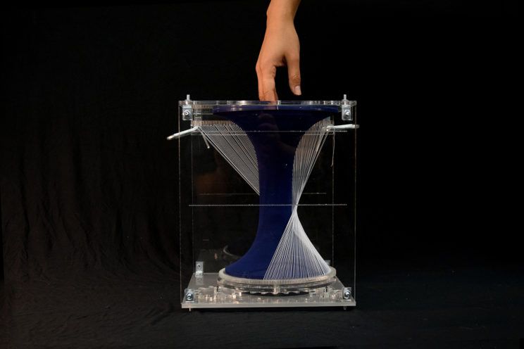 hand hovering over textile form through a hole in one face of the vertical acrylic box