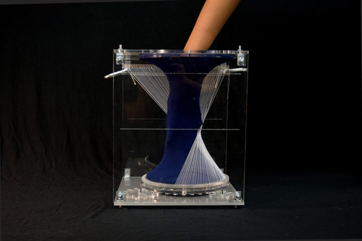 hand partially entering textile form through a hole in one face of the vertical acrylic box