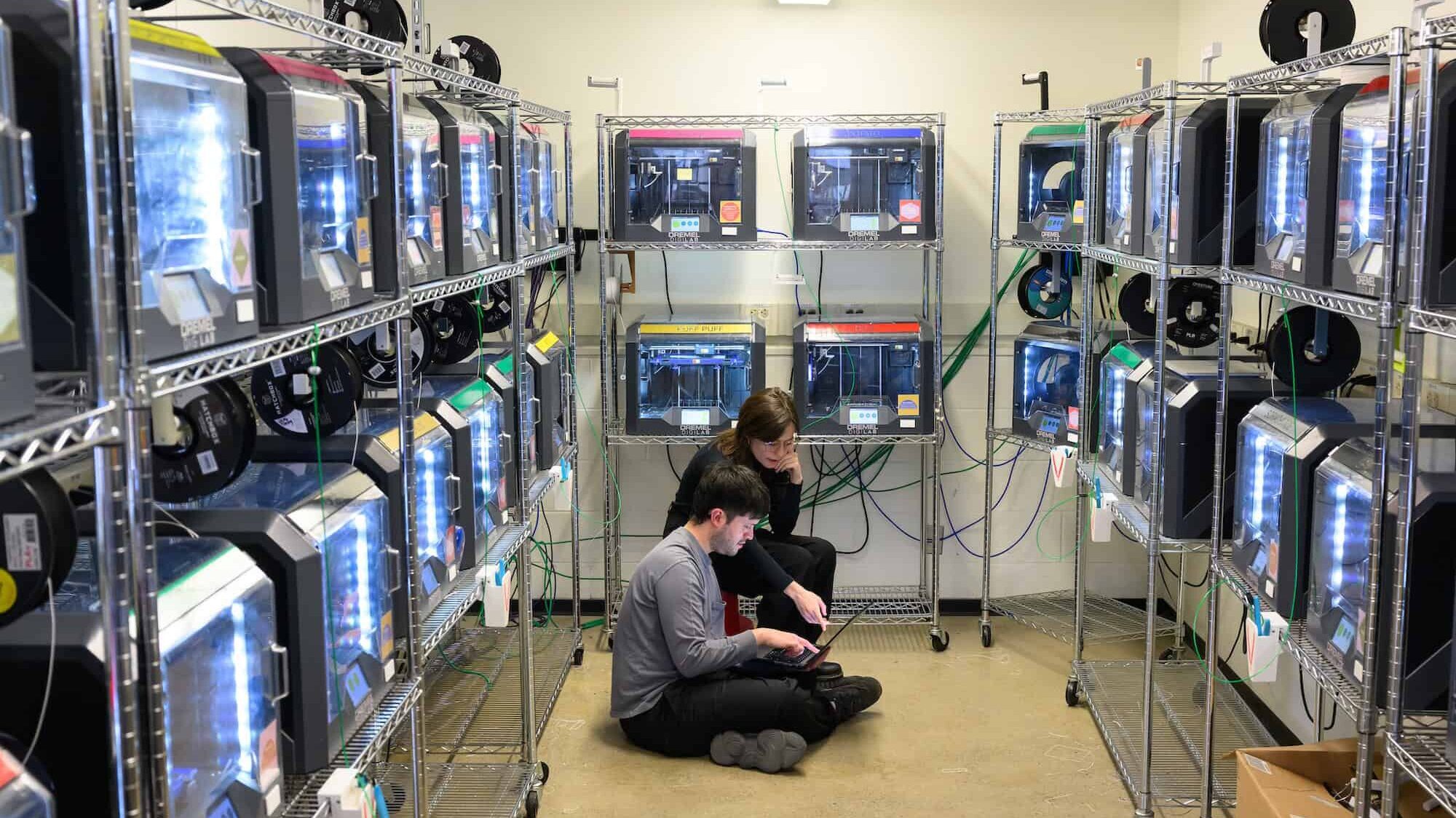 Two people sit on the floor surrounded by shelves of 3D printers.